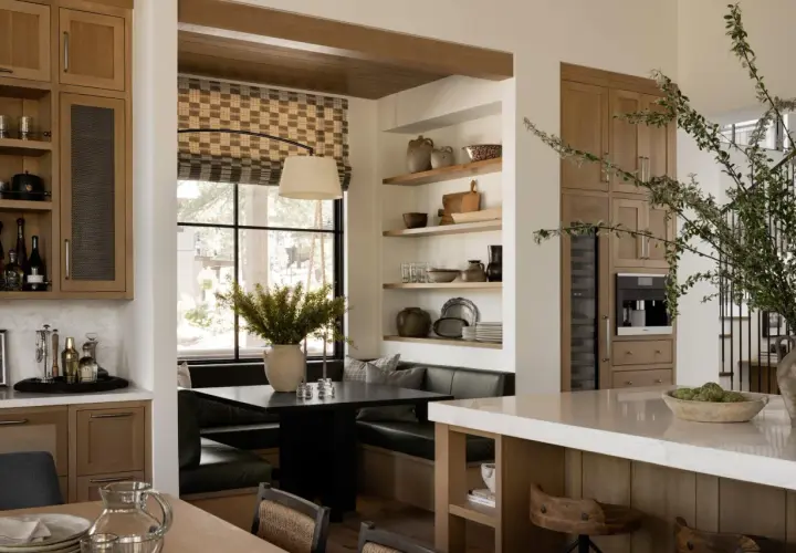 A cozy breakfast nook built in with a dark table, plush seating, and a large window. Shelves display rustic pottery and kitchenware.