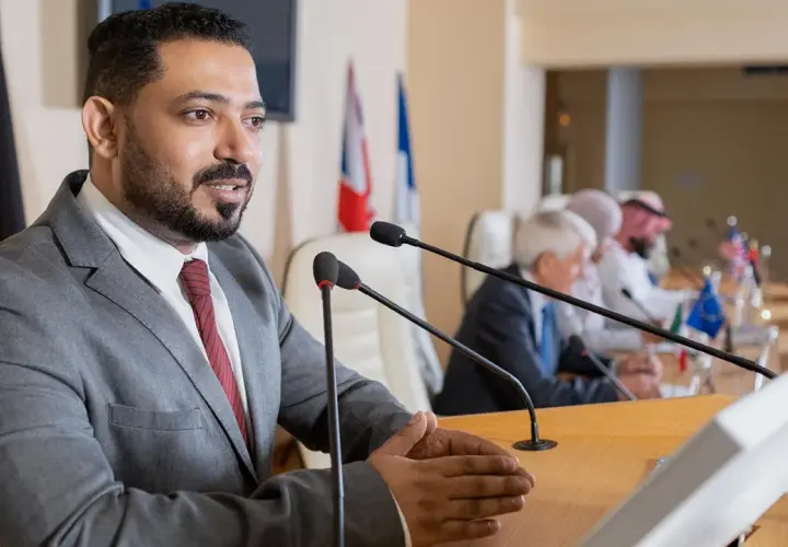 A man speaks at a podium, perhaps sharing insights from his pursuit of best online MPA programs, as others listen at a long table with flags.