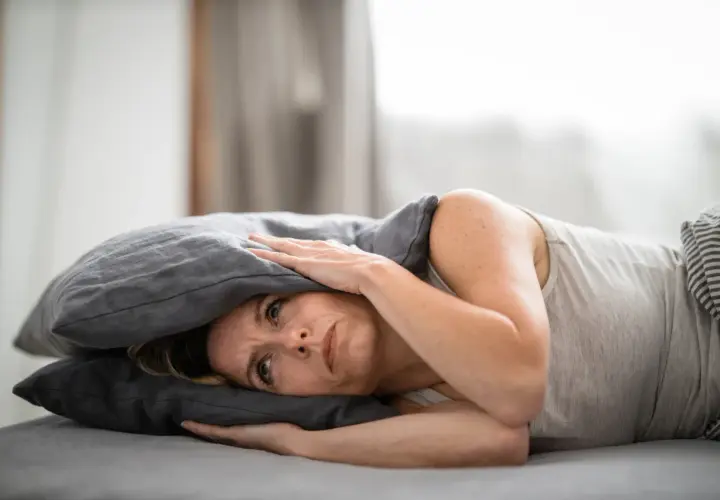 Frau mit Kissen über dem Kopf, die unter Tinnitus leidet und nach Linderung durch Osteopathie sucht.