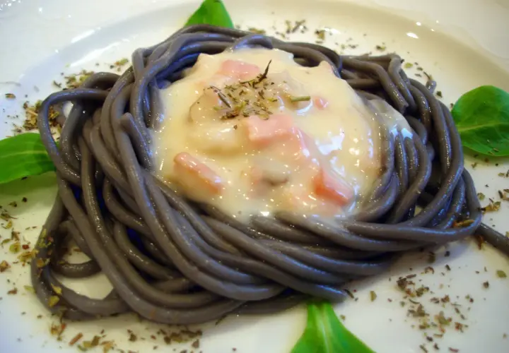 Espaguetis negro con nata, trocitos de jamón y hierbas aromáticas, servido en un plato blanco.