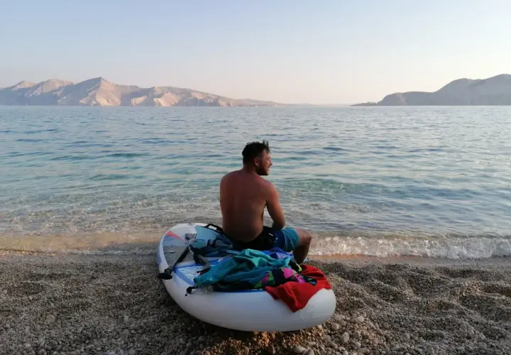 Chorwacja: mężczyzna na desce SUP na kamienistej plaży, w tle góry i spokojne morze. Idealne miejsce na relaks.