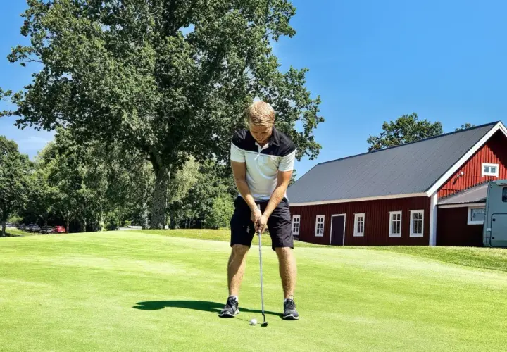 Man puttar på en golfbana, en röd byggnad och träd i bakgrunden. Golf som friskvård är en härlig aktivitet.