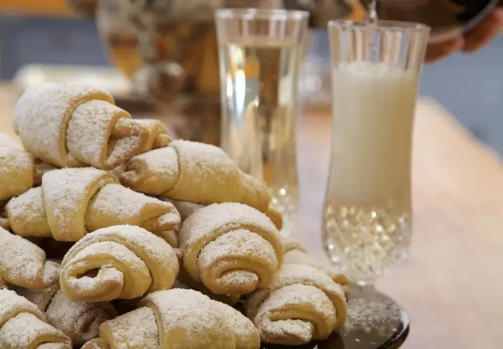 Stos puszystych rogalików krucho drożdżowych, posypanych cukrem pudrem, obok kieliszków z napojem.
