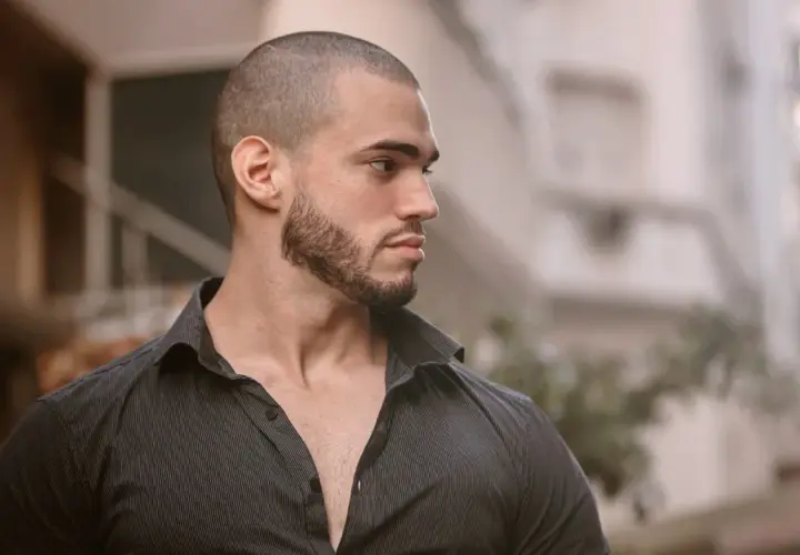 Hombre con buzz cut con entradas, barba cuidada y camisa oscura, mirando hacia un lado con expresión pensativa.