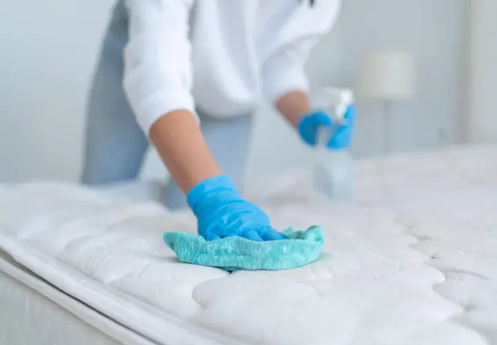 Persona con guantes azules limpia un colchón con un paño turquesa y spray. Aprende como limpiar colchones en casa para un descanso higiénico.