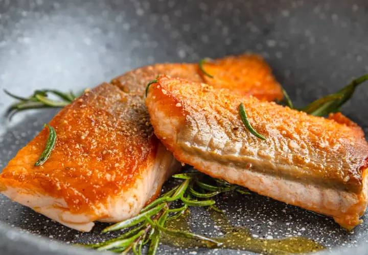 Zwei perfekt gebratene Lachsfilets mit knuspriger Haut und Rosmarin in einer Pfanne.