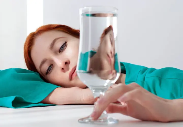 Mujer pelirroja recostada, con su rostro reflejado en un vaso de agua, quizás pensando en los beneficios de un alcalinizador de agua para su bienestar.