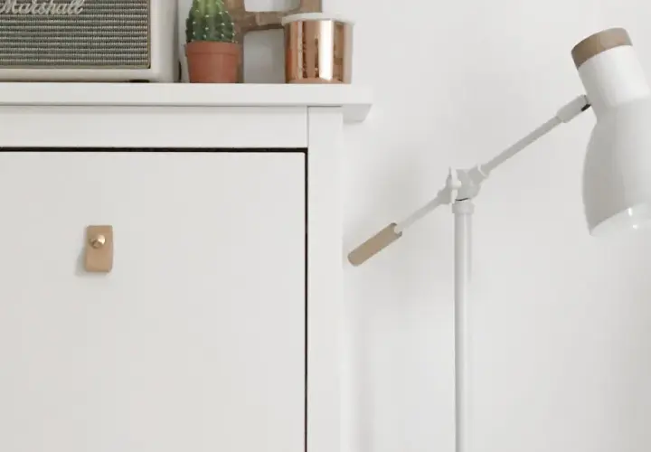 Un mueble blanco con un tirador de cuero y un altavoz Marshall. Una lámpara blanca con detalles de madera ilumina la escena.