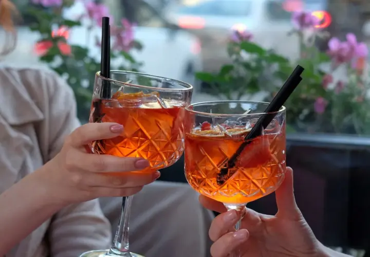 Dwie osoby wznoszą toast Aperol Spritz, ozdobionymi owocami i słomkami, na tle kwitnących kwiatów.