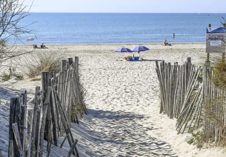 Ein sandiger Weg führt zum Montpellier Strand. Zwei Sonnenschirme spenden Schatten, während Menschen die Sonne genießen.