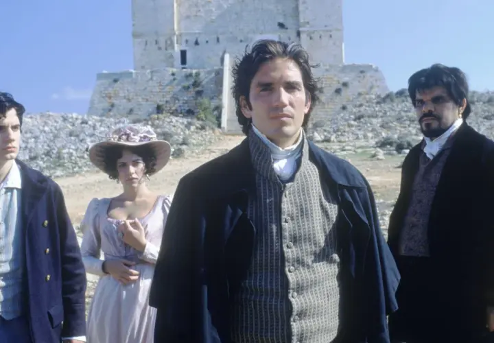 Młody mężczyzna w stroju z epoki, jak z filmu hrabia Monte Cristo 2024, stoi na tle kamiennej budowli. Obok niego kobieta w kapeluszu i inny mężczyzna.