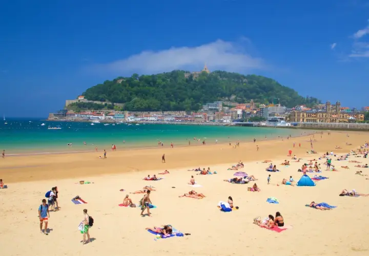 Gente disfrutando de la arena dorada y el mar turquesa en la que podría ser la mejor playa de España, con la ciudad y un monte verde de fondo.