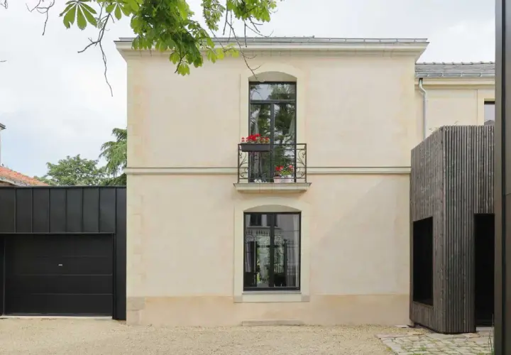 Maison rénovée par un architecte DPLG, mêlant pierre claire, bois sombre et garage noir moderne. Balcon fleuri sur la façade.