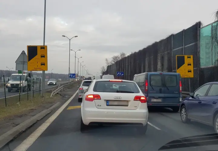 Korek na drodze, samochody stoją w miejscu. Widoczny znak "Zalecana zasada ruchu" informujący o konieczności stosowania jazdy na suwak.