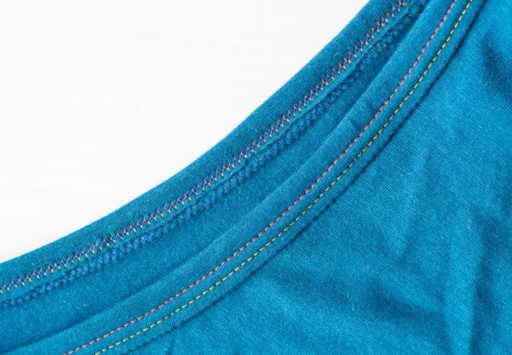 Close-up of teal fabric showing a decorative double needle topstitch with colorful threads, adding a vibrant detail to the garment's edge.