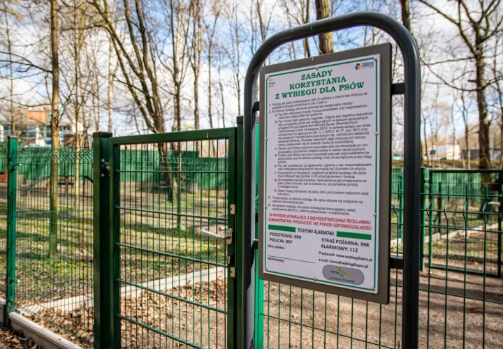 Zielona brama do parku dla psów, z tablicą informującą o zasadach korzystania.