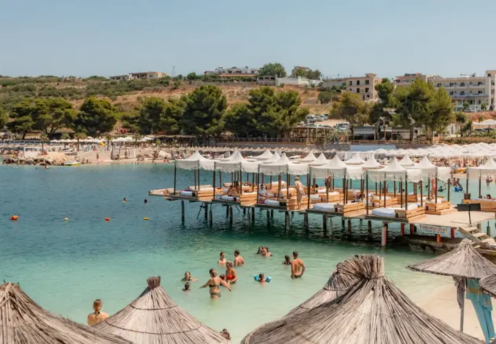 Słoneczny dzień na plaży Ksamil. Ludzie kąpią się w turkusowej wodzie, odpoczywają pod białymi parasolami na pomostach i na piaszczystym brzegu.