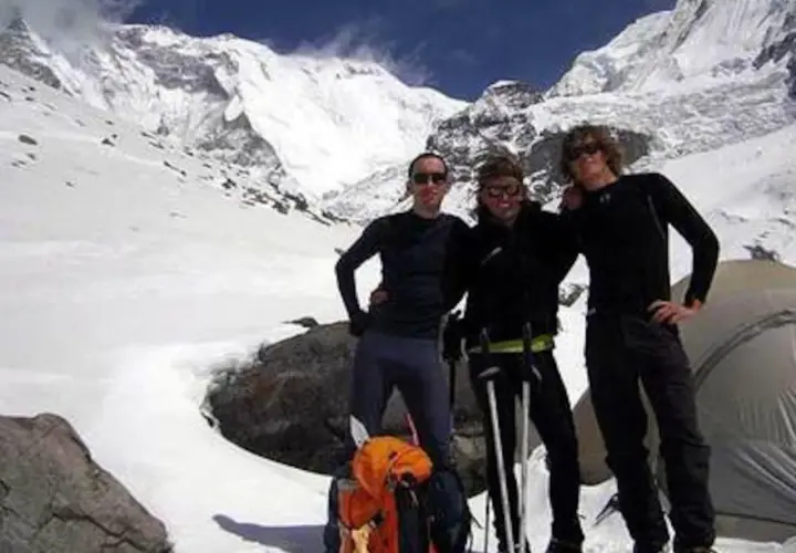 Tres montañistas posan con equipo de esquí y mochila frente a un paisaje nevado y montañoso.