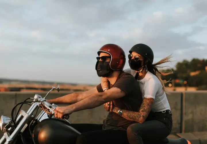Pareja con mascarillas, montando a horcajadas moto al atardecer. El viento agita el cabello de ella.
