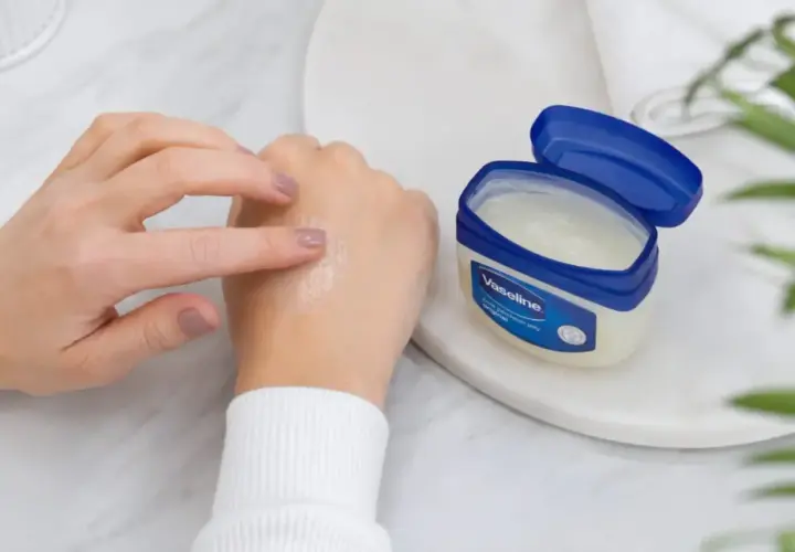 Dłoń rozsmarowuje na skórze nawilżający krem z wazeliną. Obok stoi otwarty słoiczek z produktem.