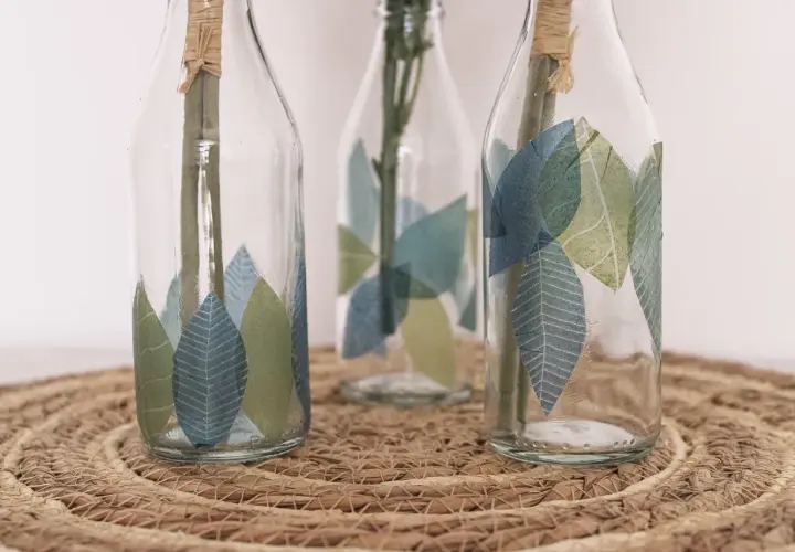 Schöne Serviettentechnik Ideen: Glasflaschen mit Blattmotiven verziert, auf einem geflochtenen Untersetzer.