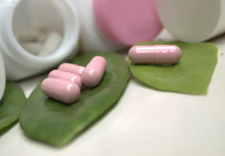 Różowe kapsułki, być może zawierające ketoprofen, leżą na zielonych liściach obok otwartych pojemników z lekami.