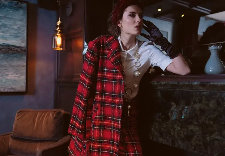 Une femme pose, vêtue d'un blazer au **motif tartan** rouge et noir, avec un collier perlé.