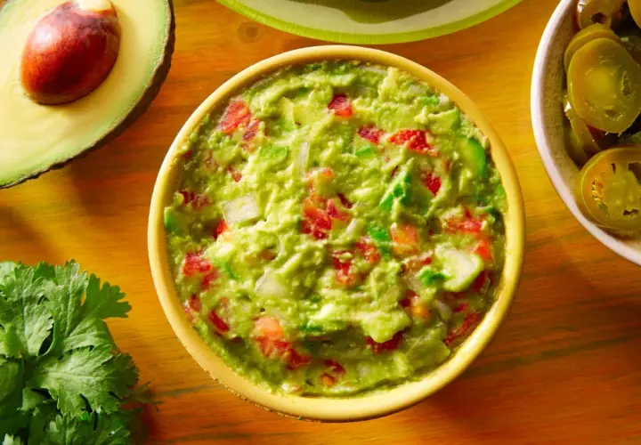 Miseczka guacamole z pomidorami i cebulą, obok awokado, kolendra i plasterki jalapeno.