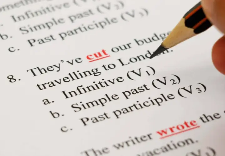 Matita evidenzia "cut" e "wrote" su un foglio con esercizi di grammatica inglese, tra cui la forma infinito.