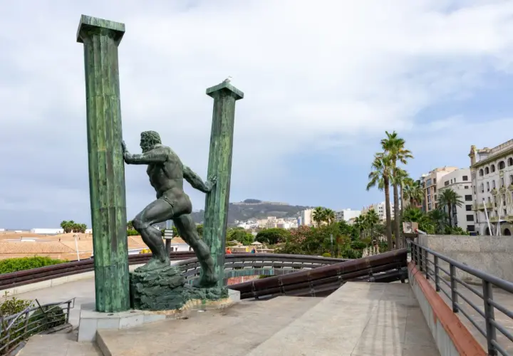 Statua di Ercole che sorregge le colonne d'Ercole oggi, con palme e edifici sullo sfondo.