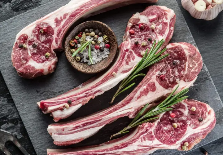 Côtelettes d'agneau crues, assaisonnées de sel et de poivre, prêtes pour une recette d'agneau pascal. Romarin frais et ail complètent la scène.