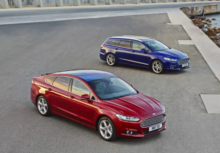 Dwa samochody Ford MK4: czerwony sedan i niebieskie kombi, stoją na asfaltowej nawierzchni przy betonowej konstrukcji i morzu.