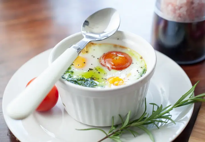 Un œuf ramequin cuit au four, garni d'une tomate et d'herbes, prêt à être dégusté avec une cuillère.