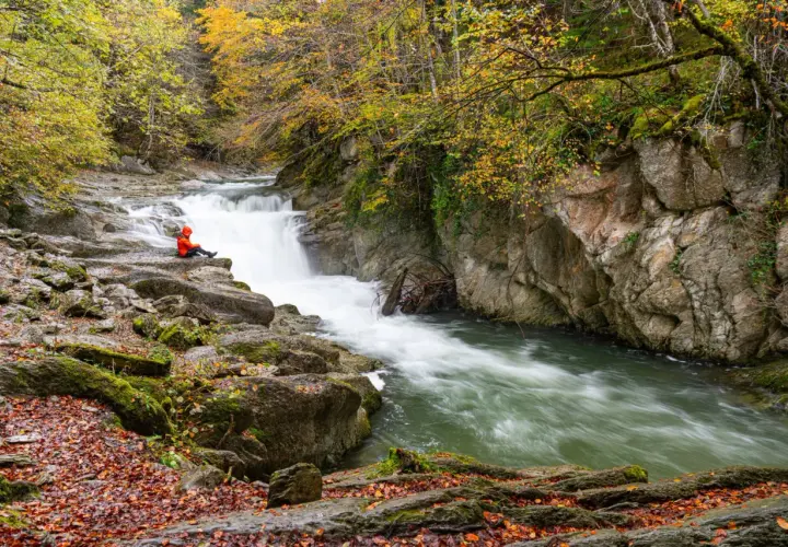 Persona sentada en rocas junto al Saut deth Pish, rodeada de árboles otoñales y agua turbulenta.