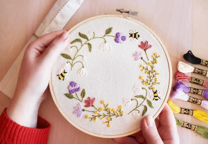 Maintenant, je peux vous aider avec ça. Voici une description alternative pour l'image :

Main tenant un tambour à broder avec des abeilles et des fleurs. Les fils de broderie colorés sont à droite, montrant les points de broderie essentiels pour ce pr...