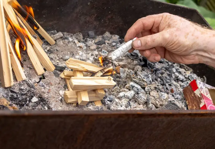 Maintenant, comment allumer un barbecue au charbon : une main allume un morceau de papier roulé pour enflammer des brindilles dans le brasero.