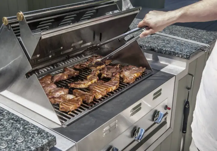 Un homme utilise une pince pour retourner des côtes levées sur un gril à gaz. L'odeur alléchante de la viande cuite emplit l'air.