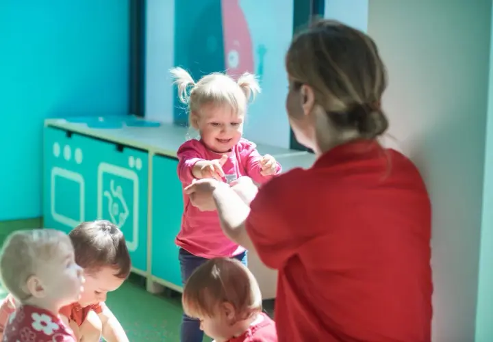 Nauczycielka w czerwonej bluzce bawi się z dziećmi w różowych i czerwonych ubrankach.
