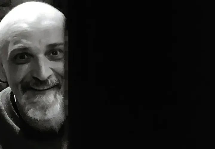 Uśmiechnięty Wojciech Sęp Dudziński, z siwą brodą i łysą głową, wygląda zza ciemnej zasłony.