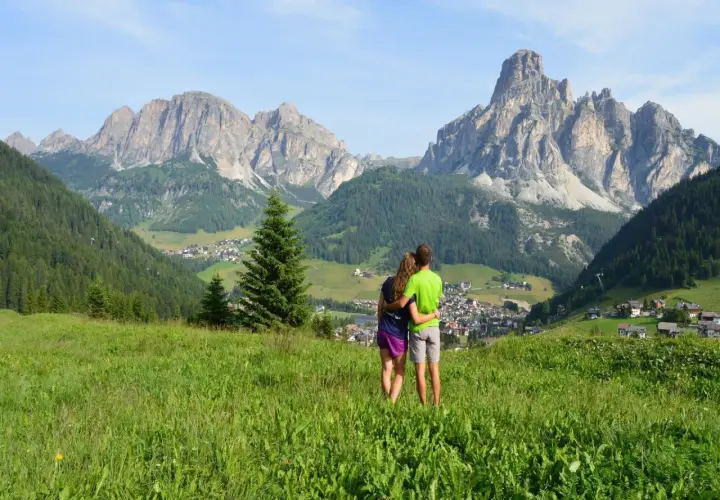 Pareja abrazada contemplando el paisaje alpino, un pueblo y montañas imponentes. Un momento perfecto de trekking en las Dolomitas.