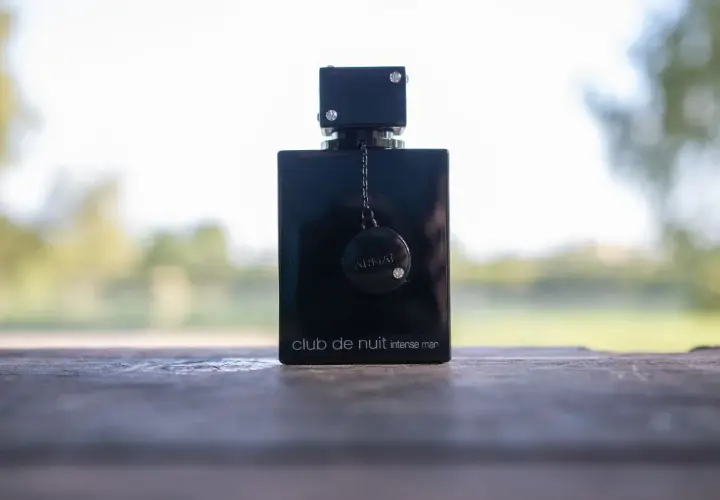 Elegancka czarna butelka perfum Club de Nuit Intense Man, ozdobiona łańcuszkiem i dyskiem z logo, stoi na drewnianym stole.
