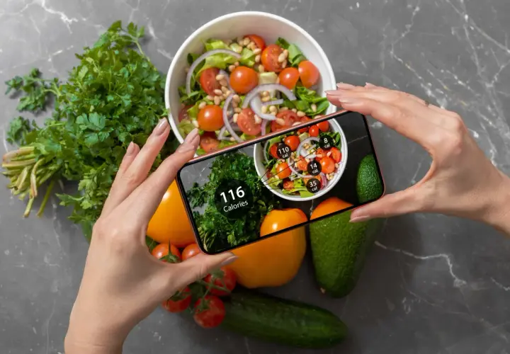 Ręce trzymają smartfon z aplikacją pokazującą kalorie. Na ekranie widać sałatkę i zioła, a obok na blacie leżą warzywa. Zastanawiasz się, ile kalorii żeby przytyć?