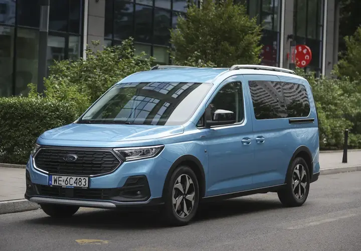 Błękitny ford 7 osobowy na tle nowoczesnego budynku. Idealny dla rodziny na wakacyjne wyprawy.