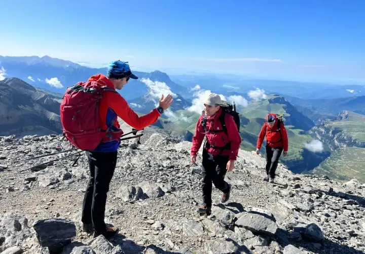 Tres excursionistas suben por una cresta rocosa en el **alt Pirineu**. El sol brilla en un cielo azul claro, con vistas a un vasto paisaje montañoso.