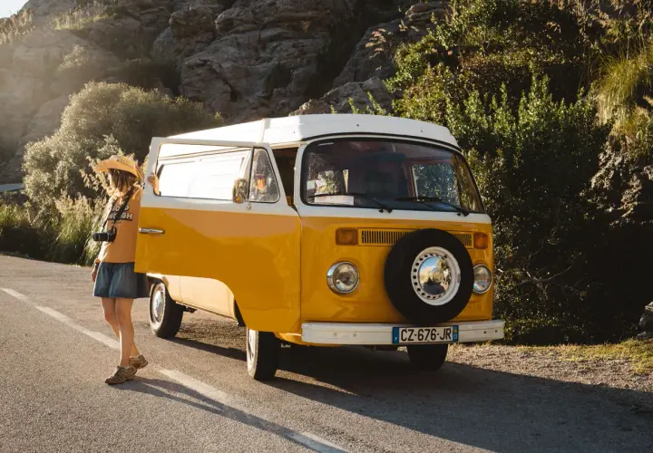A woman with a straw hat and camera leans against a yellow van, ready for an adventure in Majorque en van.