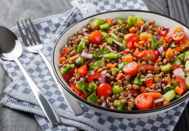 Un'insalata di lenticchie colorata e fresca, con pomodorini, peperoni, cipolla rossa e carote. Servita in una ciotola bianca, accanto a posate d'argento su un tovagliolo a quadri blu.