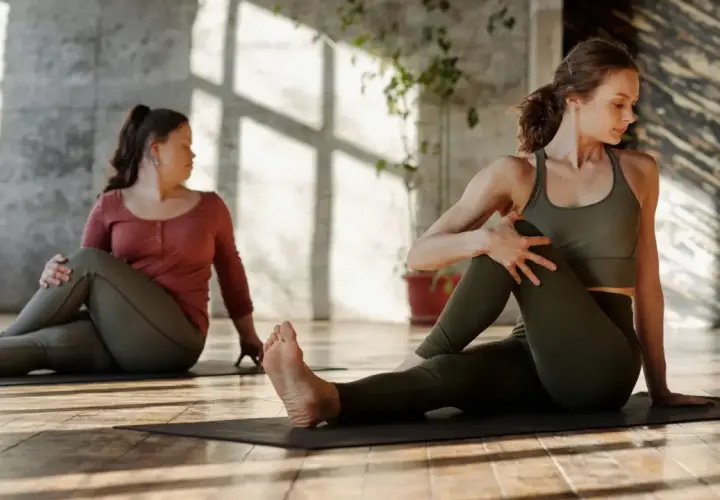 Dwie kobiety wykonują skręt tułowia na macie. To ćwiczenie to stretching, który poprawia elastyczność.