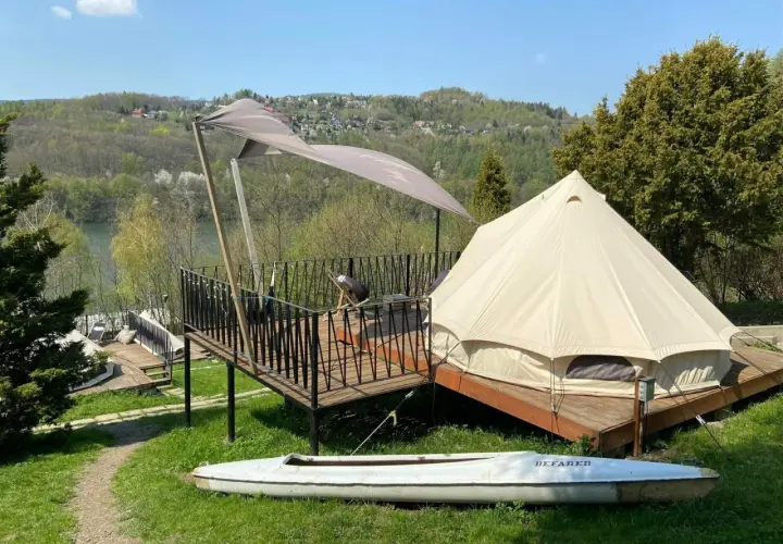 Namiot typu glamping camp z widokiem na jezioro i wzgórza. Na tarasie stolik i krzesło. Obok leży kajak.