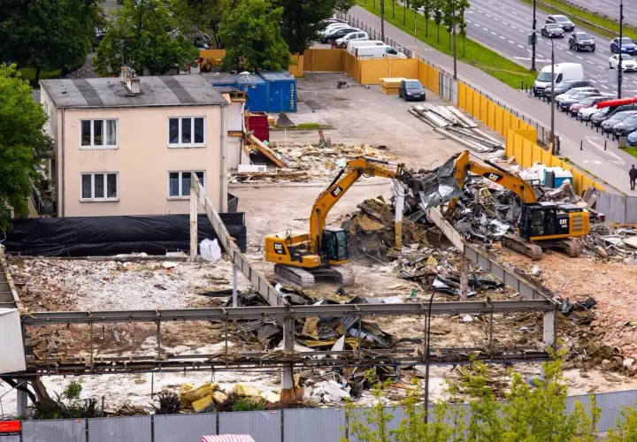 Dwa koparki pracują na placu budowy, rozbierając budynek. W tle widać drogę z samochodami i drzewa.