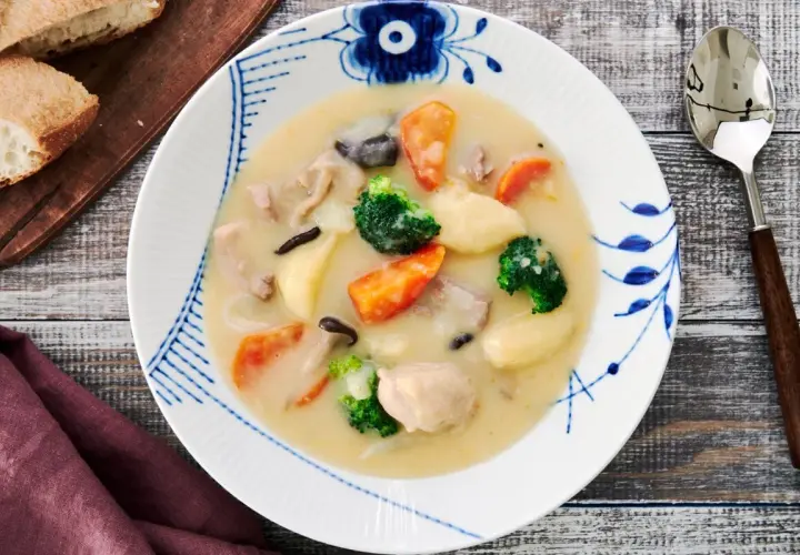 A hearty bowl of Japanese cream stew, brimming with chicken, carrots, potatoes, broccoli, and mushrooms, served with crusty bread.